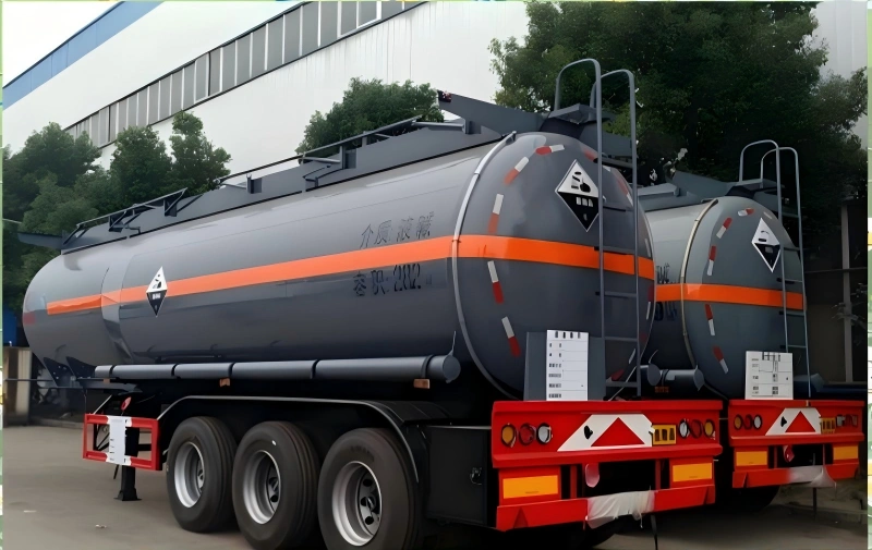 Two grey tanker trailers with orange stripes parked side-by-side outside an industrial facility. The tanks are marked with hazard warning diamonds and Chinese text specifying the medium as liquid alkali, designed for transporting corrosive substances.