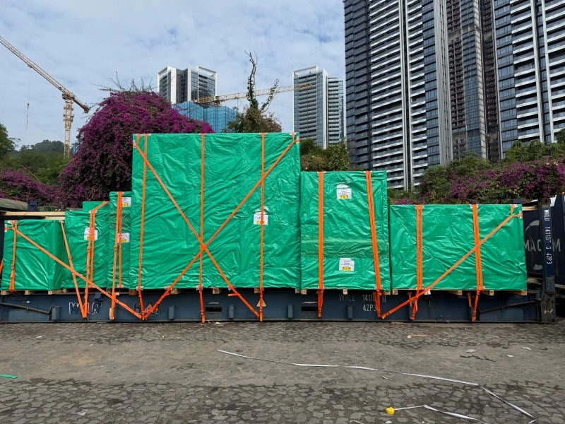 Multiple large crates, wrapped in green weather-resistant tarpaulins and secured with orange lashing straps, are loaded onto a flat rack shipping container in a port area with city skyscrapers in the background.