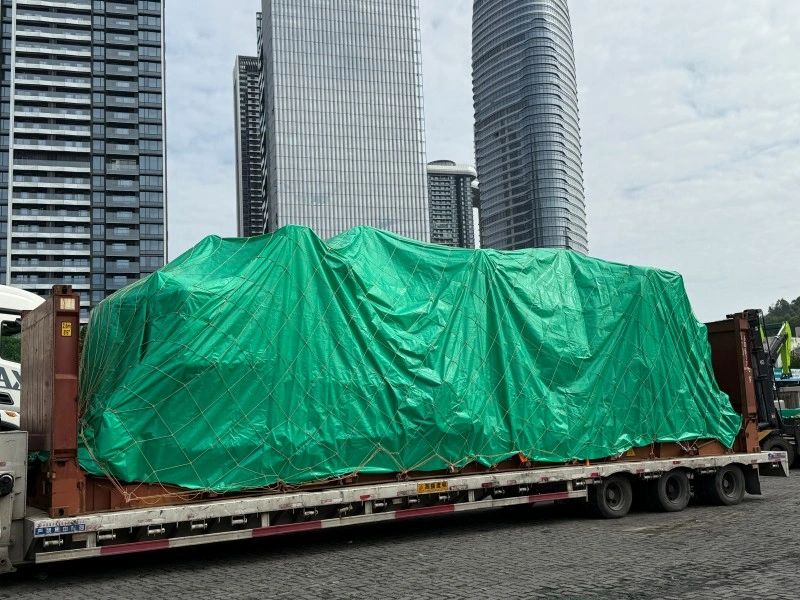 A large piece of equipment on a flat rack container is completely covered by a reinforced green tarpaulin and secured with a rope net, positioned in an urban logistics area with modern skyscrapers in the background.