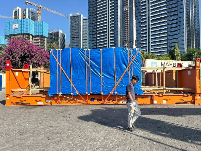A large shipment secured on an orange flat rack container is covered with a reinforced blue tarpaulin and lashing belts, demonstrating protection against rain, UV rays, and sea spray during port storage.