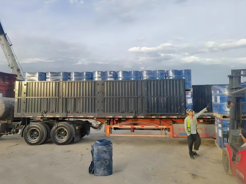 A worker directing a forklift during the loading of blue drums onto a flatbed trailer, with a crane in the background, showcasing the human element in real-time logistics management for oversized cargo.