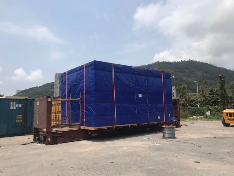 A massive piece of equipment covered in blue tarp and strapped down on an open top container, with visible wooden supports and red lashing belts, highlighting secure packing for heavy OOG shipments.