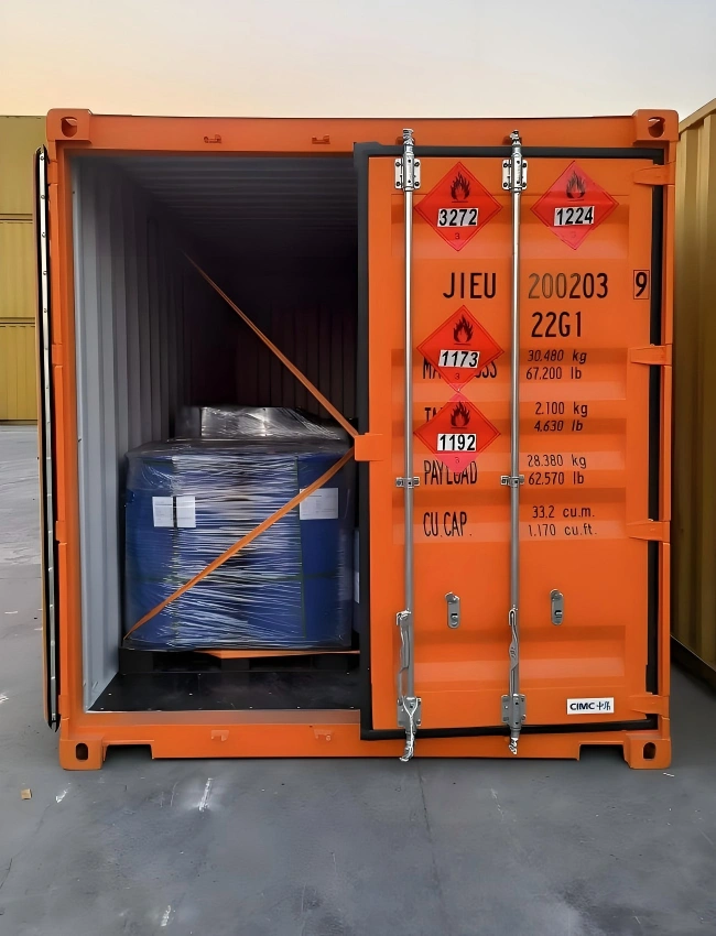 Orange DG shipping container with UN hazard class labels 3272 1192 and 1224 containing strapped blue drum for flammable liquid shipment compliant with IMDG Code