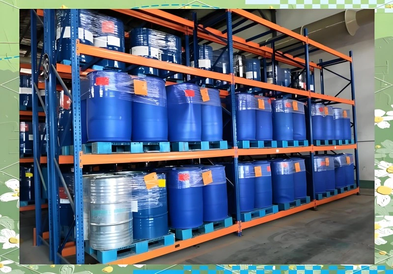 Blue and silver chemical drums neatly arranged on orange and blue pallet racking in a warehouse. The drums are shrink-wrapped on pallets, with visible hazard labels and tags, demonstrating proper chemical storage practices.