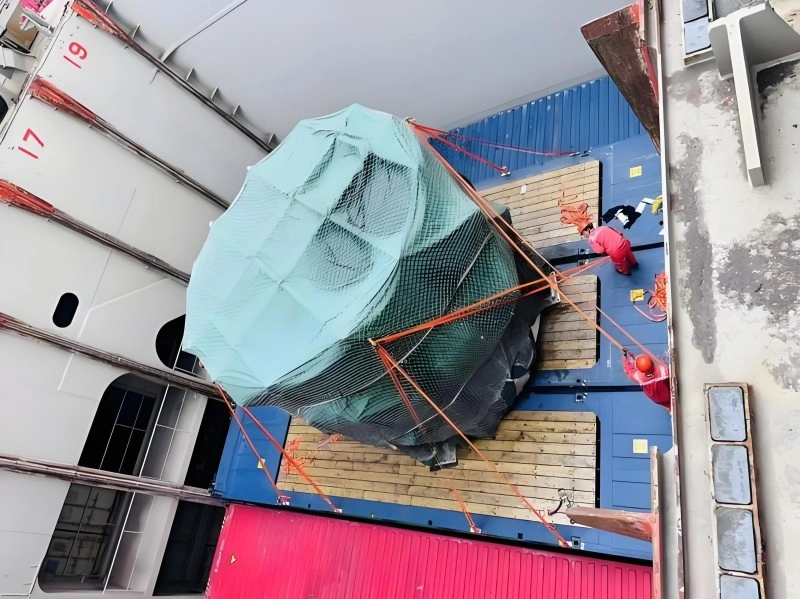 An aerial view of a large, cylindrical piece of industrial equipment, covered in green protective netting and shrink wrap, being carefully lowered by orange lifting slings onto the deck of a cargo ship, with workers in red uniforms supervising the operation.