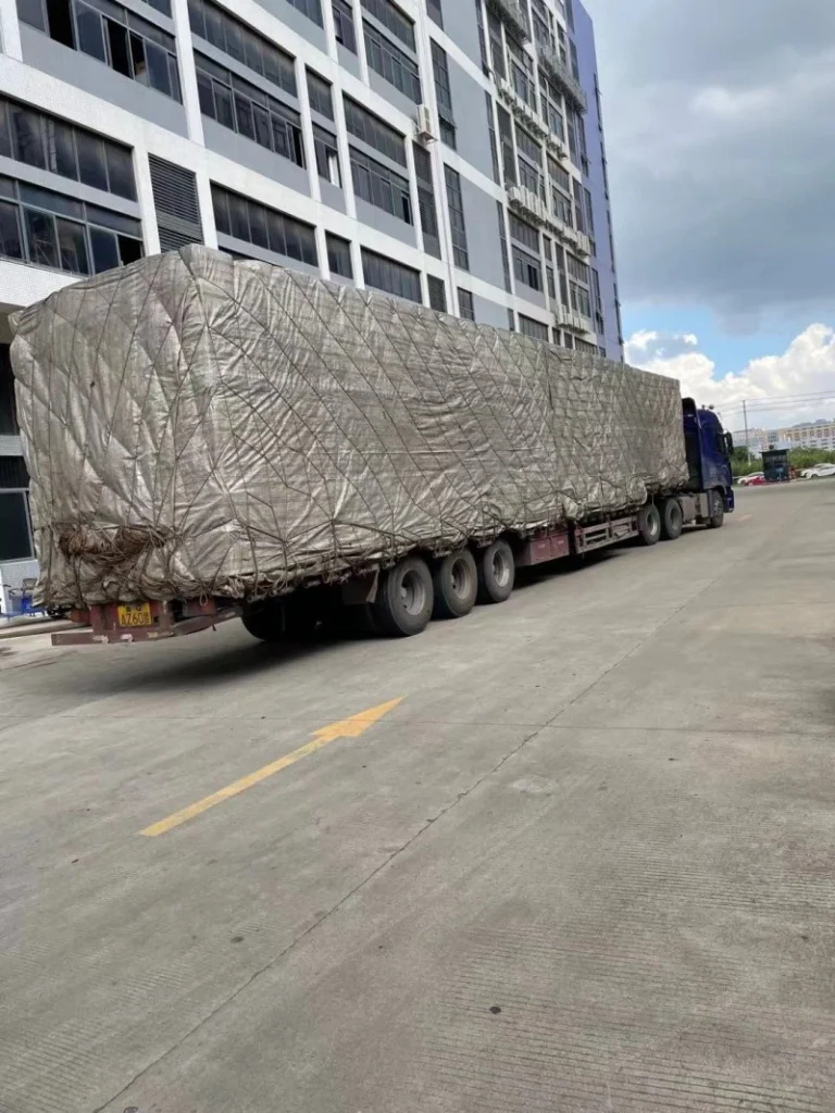 An extra-long and wide cargo unit completely covered with weatherproof tarpaulin is secured on a lowboy trailer, illustrating comprehensive protection for OOG road transportation.