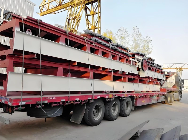 A massive, multi-section steel structure is loaded onto a red lowboy trailer, secured with multiple tie-down straps, ready for road transport in an industrial area with cranes visible in the background.