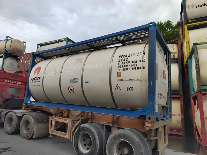 A close-up view of a white ISO tank container with blue framing, branded with the 'PEACOCK' logo. The tank displays hazard markings including 'HOT' and '115 C', along with identification numbers 'PCVU 259129 4' and '22K2'. The container is mounted on a multi-axle trailer in a container yard.