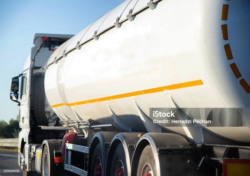 A white semi-truck with a large cylindrical pneumatic tanker trailer driving on a highway. This type of trailer is used for transporting dry bulk materials like cement or flour and features pressure relief valves and discharge systems.