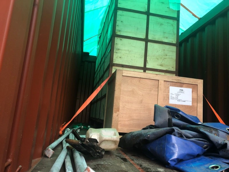 Interior view of a shipping container showing a wooden crate labeled 'PMS' and a large green-wrapped unit, secured with orange straps and blocking materials to prevent movement during transit.