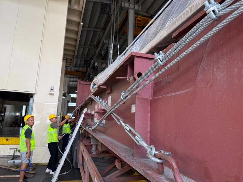Workers tightening wire rope lashings and turnbuckles to secure a large red steel structure onto a flat rack container for ocean transport.