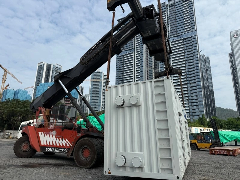 A red ContStacker reach stacker lifting a white containerized industrial unit in a logistics yard, with high-rise buildings in the background.