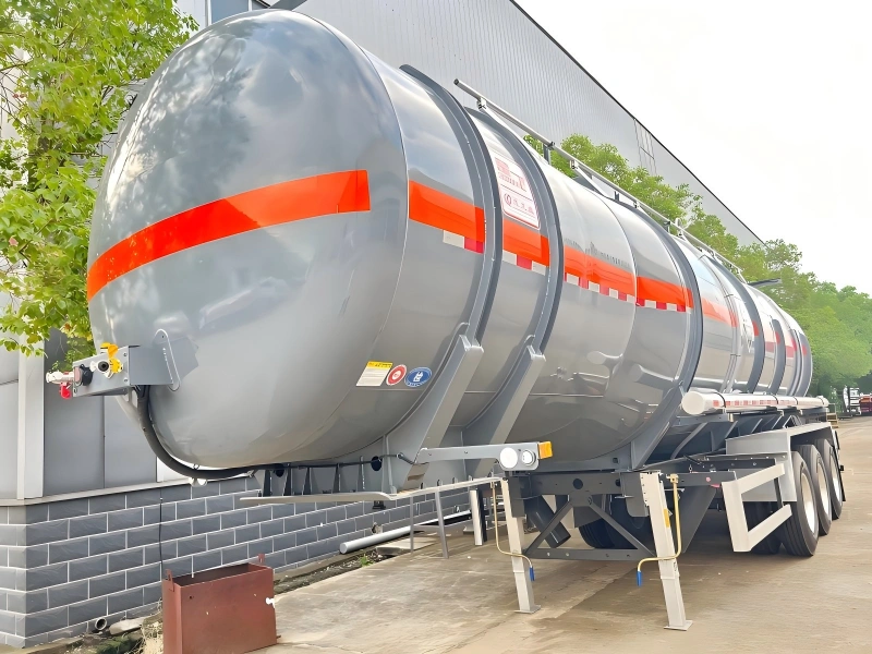 Grey road tanker equipped with orange hazard warning band and safety placards mounted on multi axle trailer for flammable or corrosive liquid transport