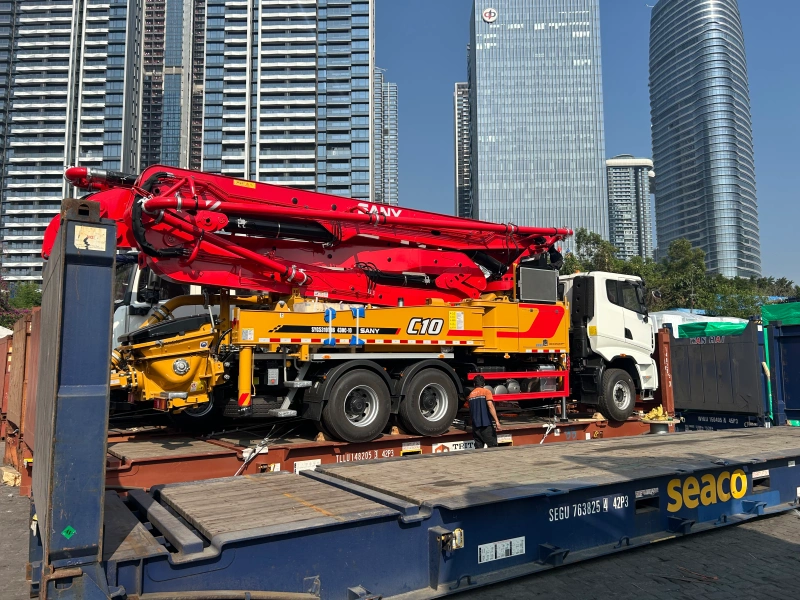 A red and yellow SANY C10 concrete pump truck is securely loaded on a flat rack container in a port terminal, with a worker inspecting the lashing, demonstrating OOG cargo preparation for international shipping.