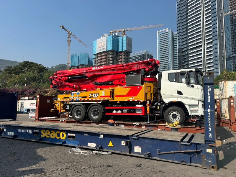 A red and yellow SANY C10 concrete pump truck is stowed on a blue flat rack container in a port terminal, exposed to open air, highlighting the need for corrosion protection against salt spray and humidity.