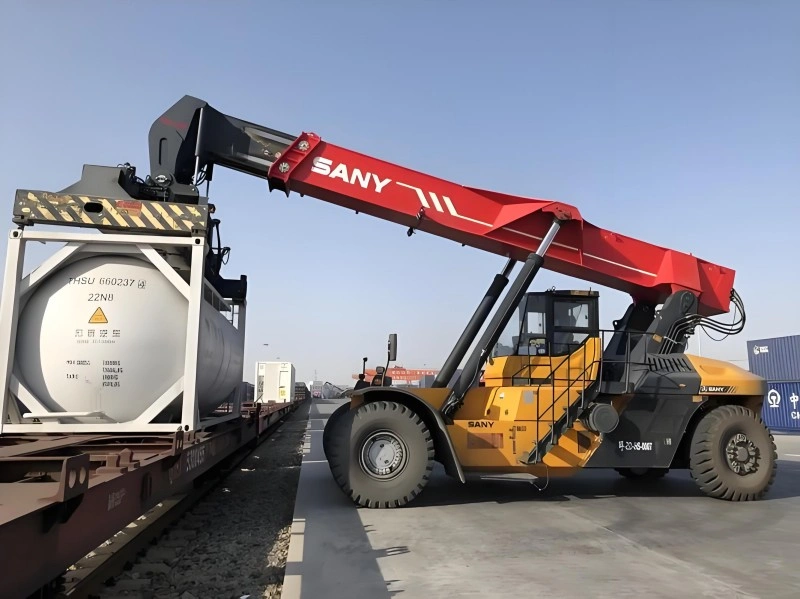 A yellow and red SANY reach stacker is carefully lifting a white tank container from a flatbed railcar at a modern rail terminal under a clear blue sky.