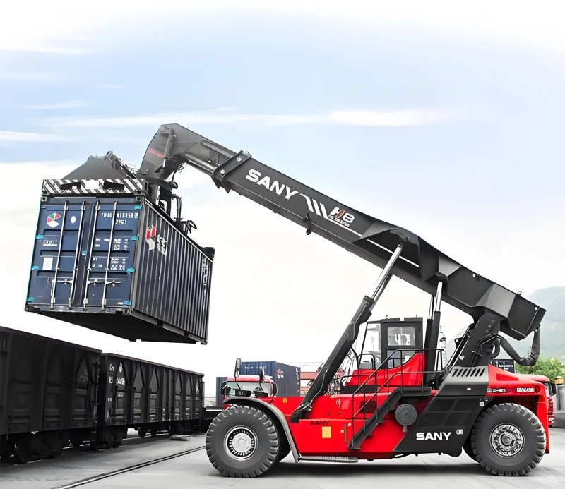 A red and black SANY reach stacker with a telescopic boom is lifting a blue shipping container and positioning it onto a freight train car in a logistics yard.