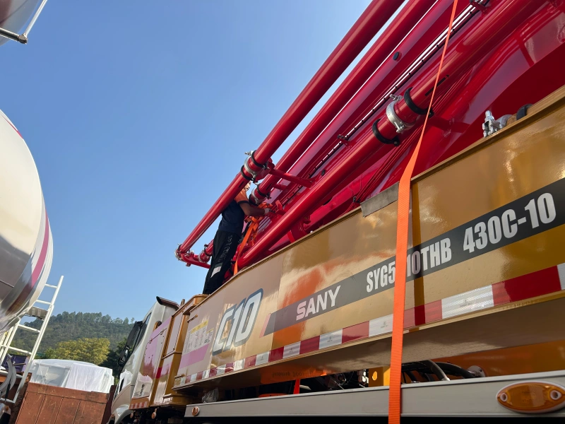 A close-up view of a worker securing the boom of a red SANY SY650THB 430C-10 concrete pump truck with orange lashing belts, highlighting the importance of proper securing for OOG cargo transport.