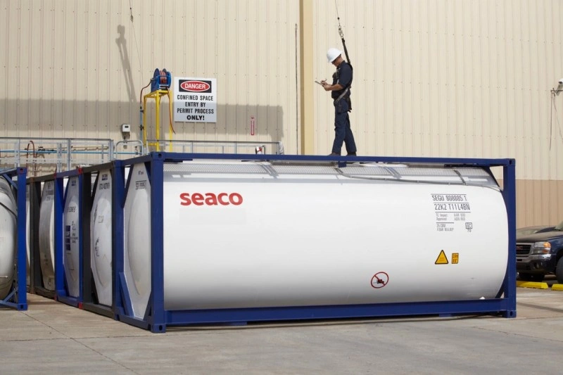 White Seaco-branded baffled liquid tank container housed in a blue ISO frame, with a worker inspecting it near a "DANGER CONFINED SPACE" sign, indicating industrial chemical handling
