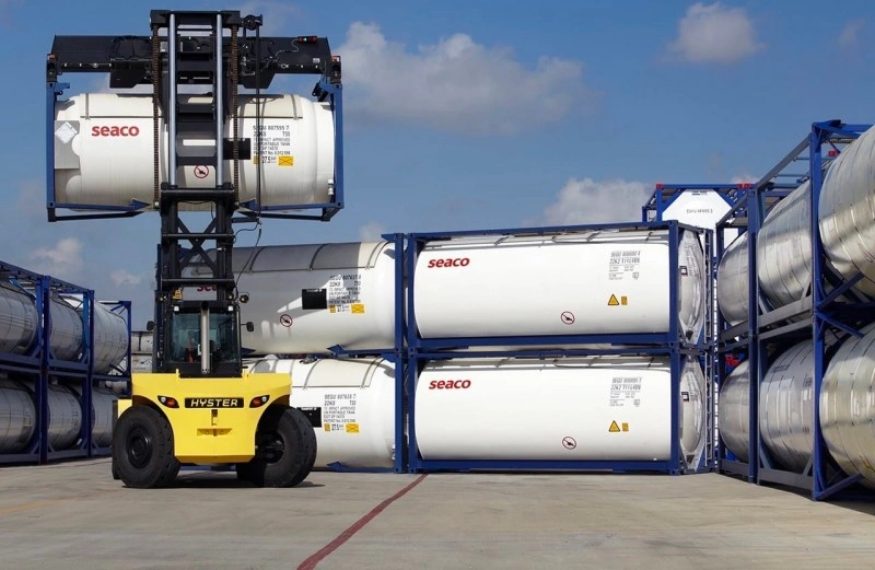 Yellow Hyster forklift lifting a white Seaco ISO tank container in a logistics yard, with multiple stacked tank containers visible in the background under clear skies