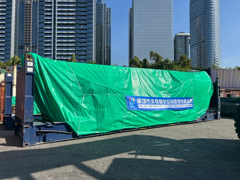An oversized cargo unit on a flat rack is fully enclosed in a green weatherproof shrink wrap or tarp, featuring a banner for "Shenzhen Bentlee Supply Chain Management," ensuring protection against condensation and weather elements.