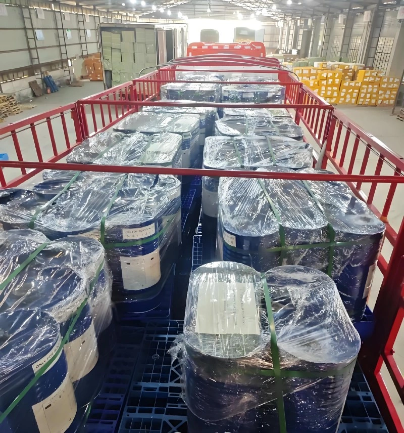 Interior view of a warehouse showing a red metal transport cart filled with blue chemical drums. The drums are stacked on blue plastic pallets and secured with clear plastic wrap and green strapping bands. Yellow storage racks are visible in the background.