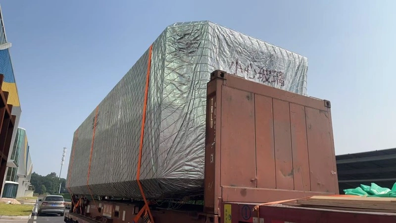 A large, silver-wrapped oversized cargo with "Handle with Care - Glass" markings, secured by orange straps on a flatbed trailer, ready for specialized logistics.