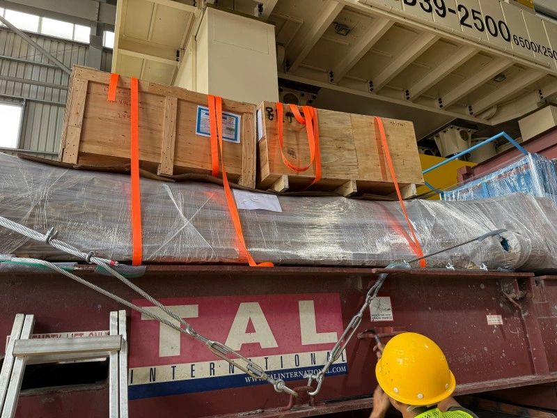 Heavy machinery wrapped in protective film and secured with orange straps on a TAL flat rack container, with a worker overseeing the loading process.