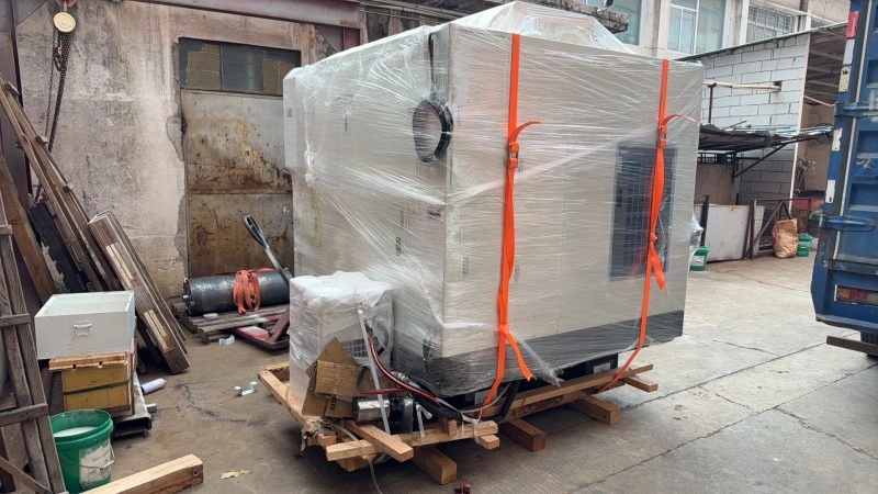 A white CNC machine fully wrapped in clear stretch film and strapped with orange heavy-duty lashing belts on a wooden skid base.