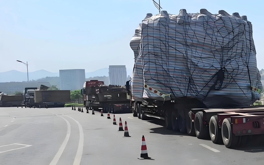 Oversized mining equipment wrapped in striped shrink film and tension netting secured on red multi axle heavy haul trailer for long haul transport