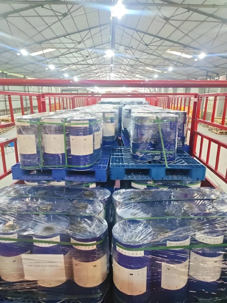 Wide shot inside a large warehouse with a high ceiling and metal truss structure. Multiple blue plastic pallets are stacked with blue chemical drums, all wrapped in clear plastic and secured with green strapping. The organized rows indicate preparation for distribution or storage.