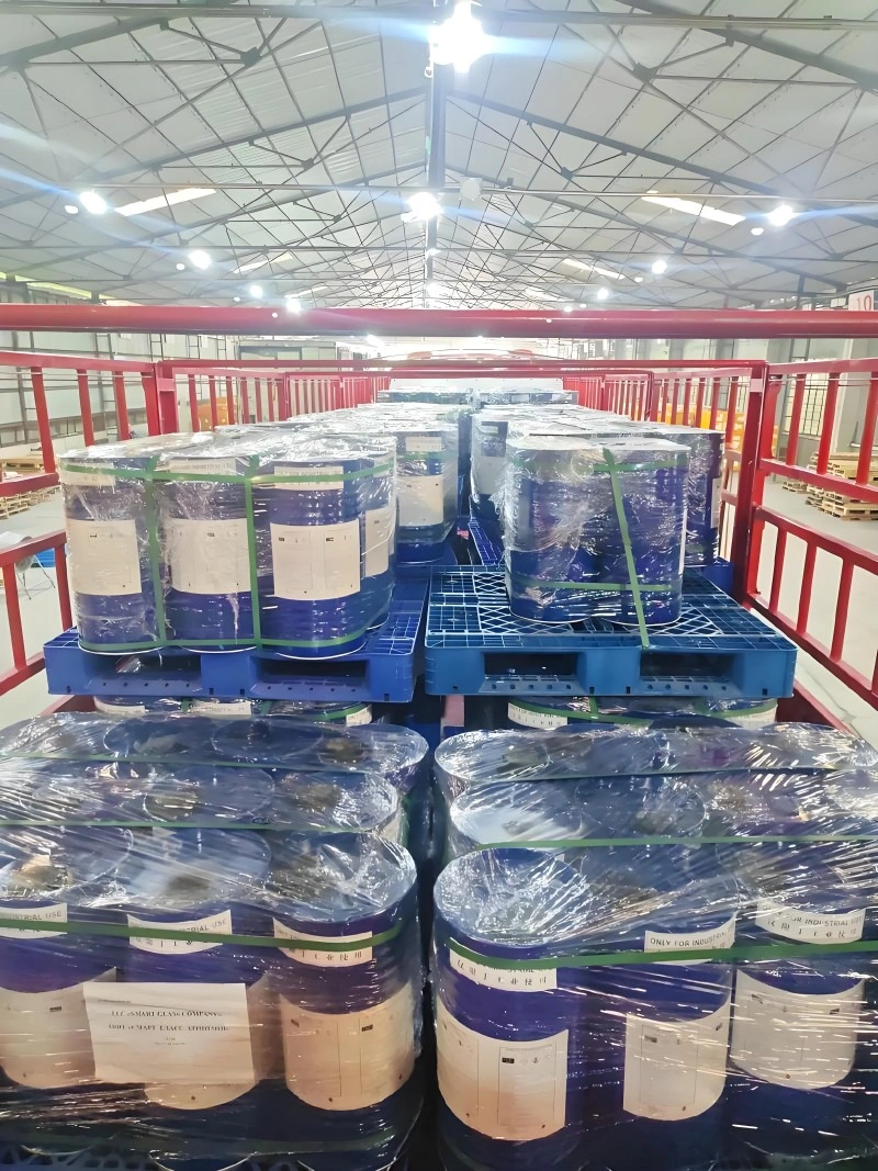 Wide shot inside a large warehouse with a high ceiling and metal truss structure. Multiple blue plastic pallets are stacked with blue chemical drums, all wrapped in clear plastic and secured with green strapping. The organized rows indicate preparation for distribution or storage.