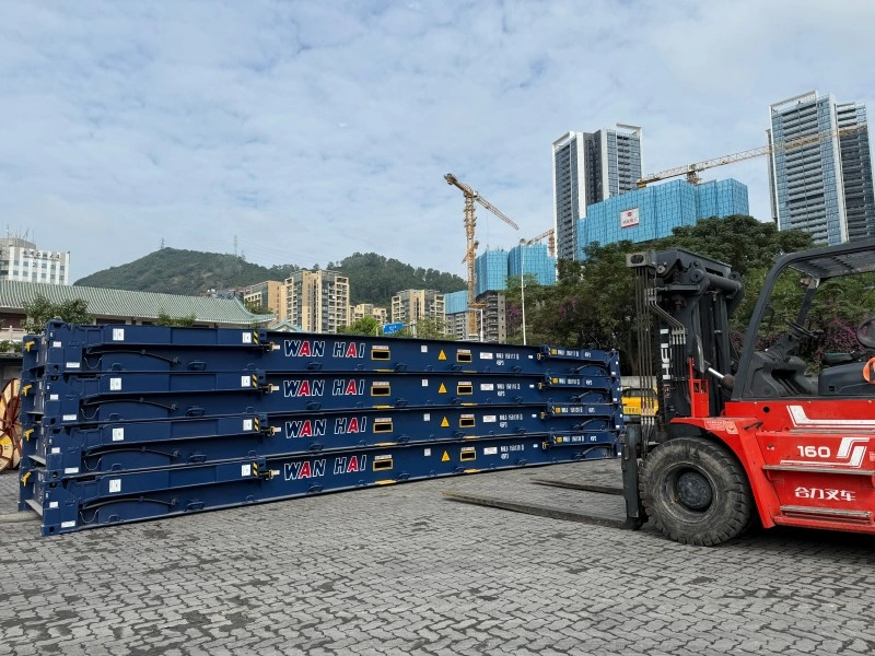 A stack of blue Wan Hai flat rack containers sits in a port yard, ready for loading oversized or heavy-lift cargo. A red forklift is positioned nearby, indicating active logistics operations for international shipping.
