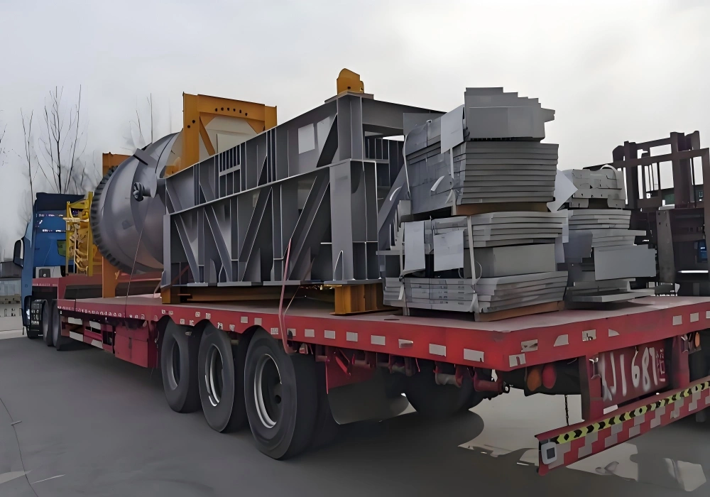 Large steel fabrication frame cylindrical tank and stacked metal plates loaded on red flatbed trailer for industrial plant equipment transport