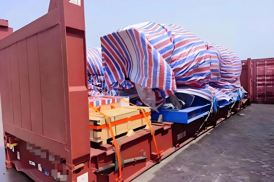 Red flat rack carrying irregular shaped cargo under striped weatherproof tarp with smaller packed units secured at front for mixed load project shipment