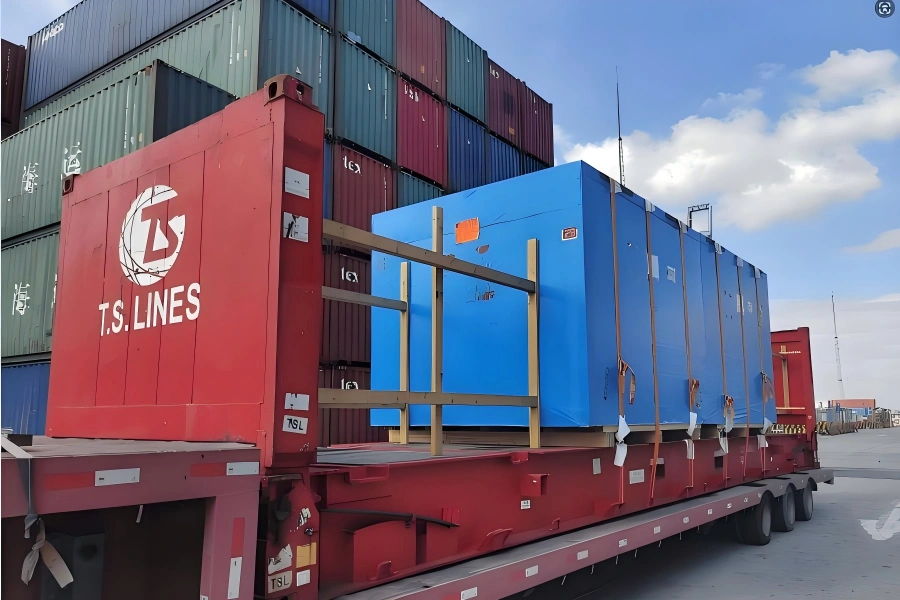 T.S. LINES branded container chassis transporting blue wrapped industrial modules on flat rack with stacked shipping containers in port yard