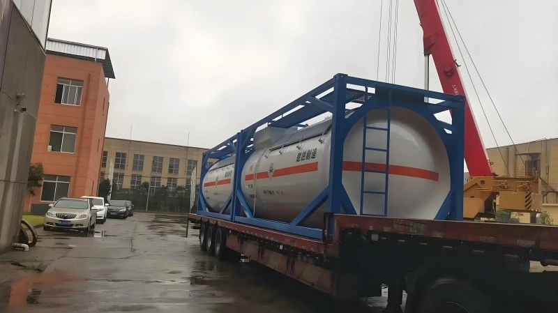 A blue-framed ISO tank container, housing two large white cylindrical tanks with orange hazard stripes, is secured on a red flatbed trailer. A red mobile crane is visible in the background, with industrial buildings and parked cars nearby on a wet, overcast day.