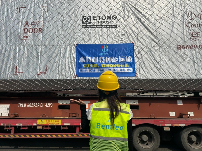A logistics supervisor inspecting a prefabricated Etong House module wrapped in protective material and secured on a heavy haul trailer.