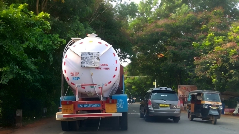 The rear view of a white vacuum tanker truck driving on a tree-lined road. The tanker is equipped with a pressure/vacuum relief valve and markings for a "BREAK SYSTEM," indicating its use in liquid waste or sewage transportation.