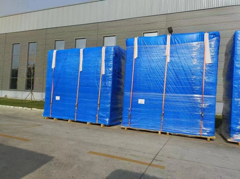 Several large cargo pallets wrapped in protective blue tarpaulin and clear stretch film, secured with orange straps, standing outside a modern industrial building.