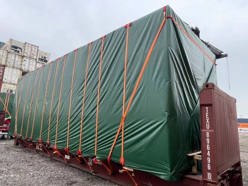 A large, rectangular piece of industrial equipment, completely wrapped in a green waterproof tarpaulin and secured with multiple orange lashing straps, loaded onto a flat rack container at a port.