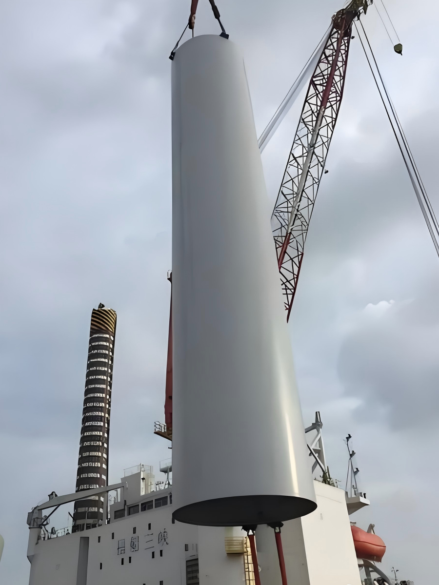 White wind turbine tower section suspended by crane during loading onto vessel deck for renewable energy project shipment