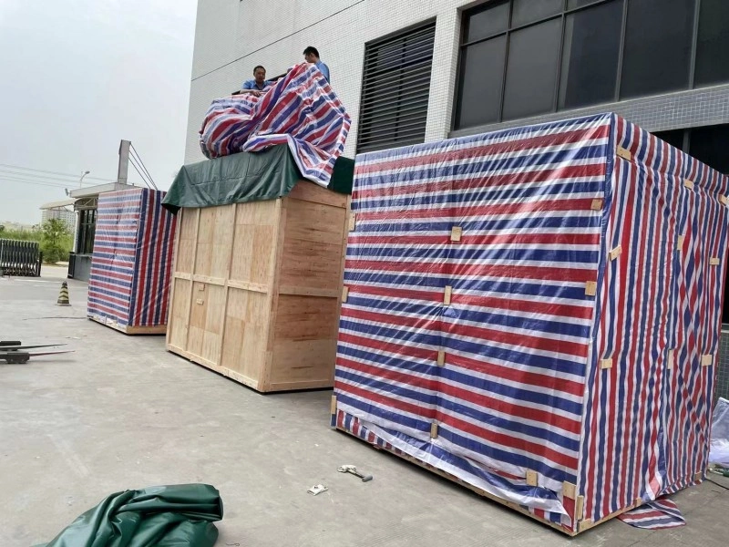 Two workers on top of a large wooden crate are covering it with a red, white, and blue striped tarpaulin, with other similarly covered crates nearby in an outdoor yard.