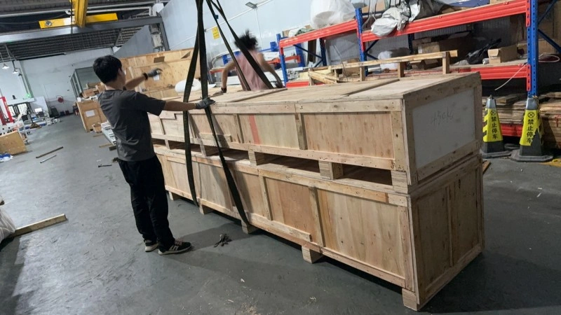 Two workers in a warehouse are using heavy-duty lifting straps to secure a large, reinforced wooden export crate containing industrial machinery, preparing it for safe handling and international shipping.