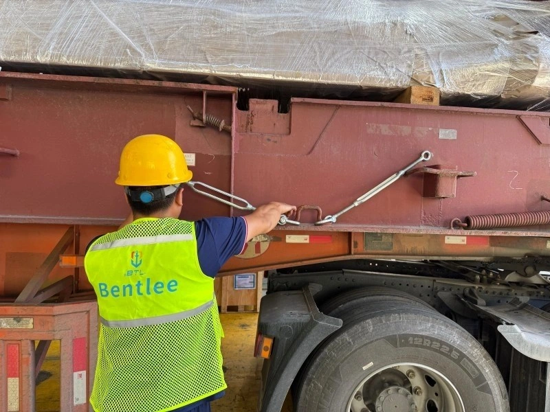 Worker in yellow hard hat and Bentlee safety vest adjusting a turnbuckle on a steel wire rope to secure a large, plastic-wrapped industrial machine on a red flatbed trailer