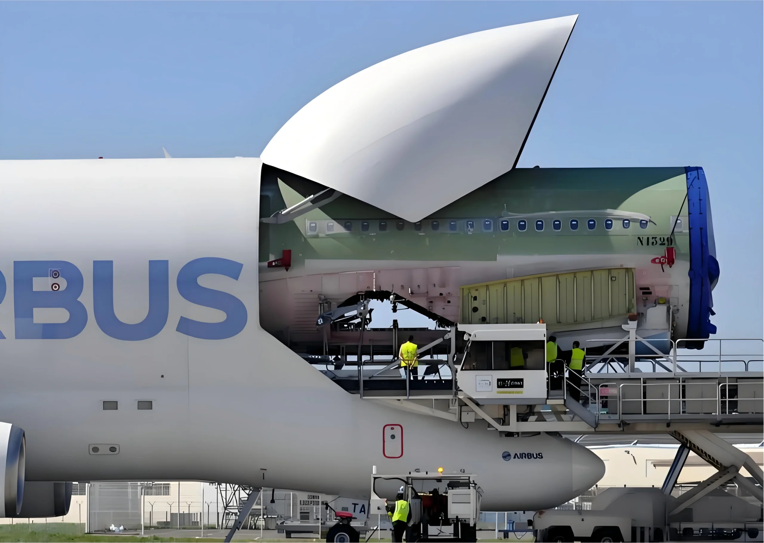 Workers in yellow safety vests operate a mobile lift platform beside the open upper fuselage of an Airbus Beluga super transporter, revealing a green-painted aircraft section inside, with clear blue skies overhead.