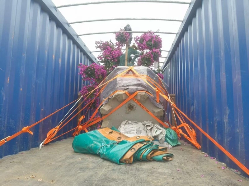 A large potted bougainvillea plant with vibrant purple flowers is loaded into a blue open-top shipping container, secured with orange lashing straps and partially covered with a gray tarp, with a worker visible in the background.