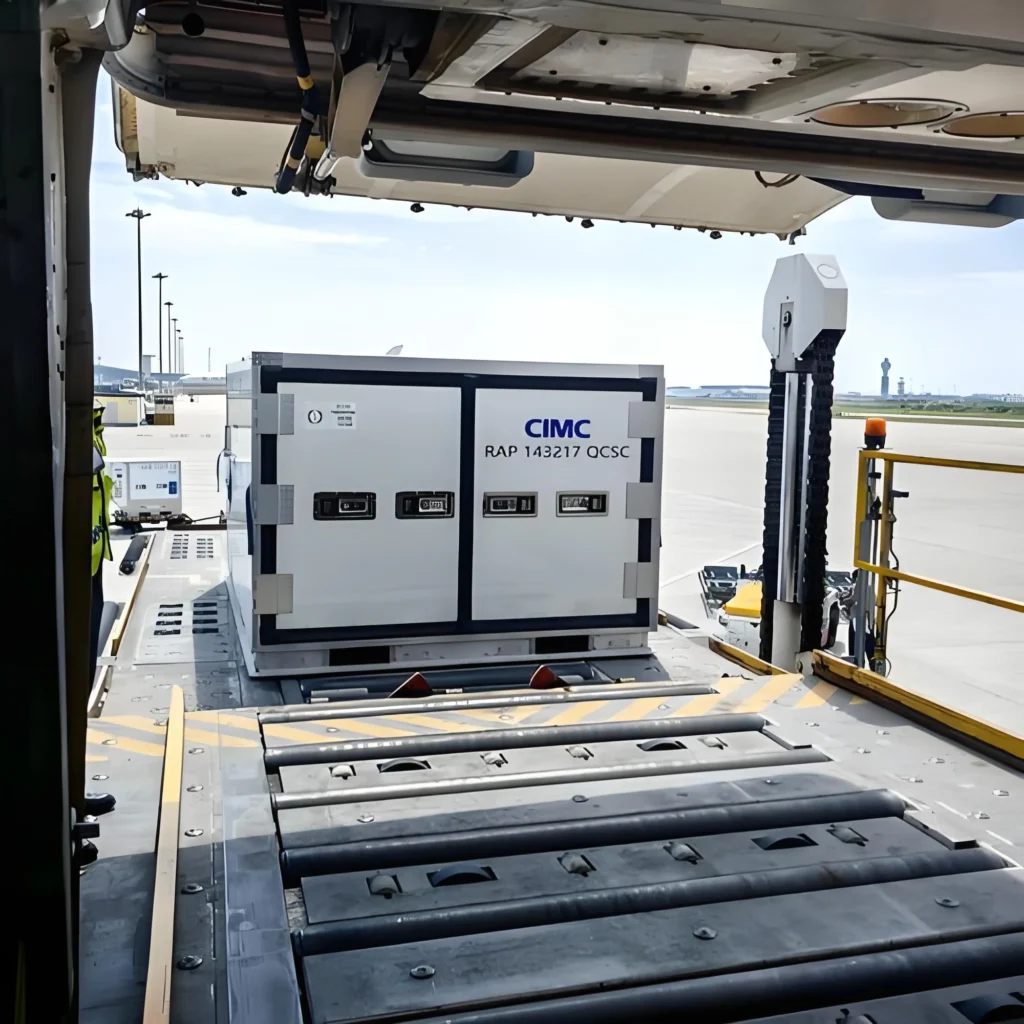 A white CIMC-branded RAP active temperature-controlled air cargo container, labeled “RAP 143217 QCSC”, is being loaded onto an aircraft via a roller conveyor system at an airport tarmac, with ground crew in high-visibility vests overseeing the operation.