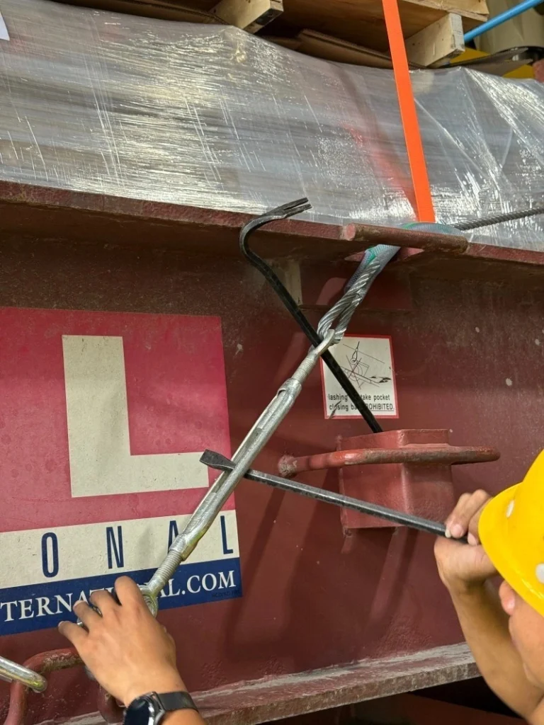 A close-up view of a worker's hands tightening a metal turnbuckle connected to a steel wire rope, anchored to the side of a shipping container or truck bed.
