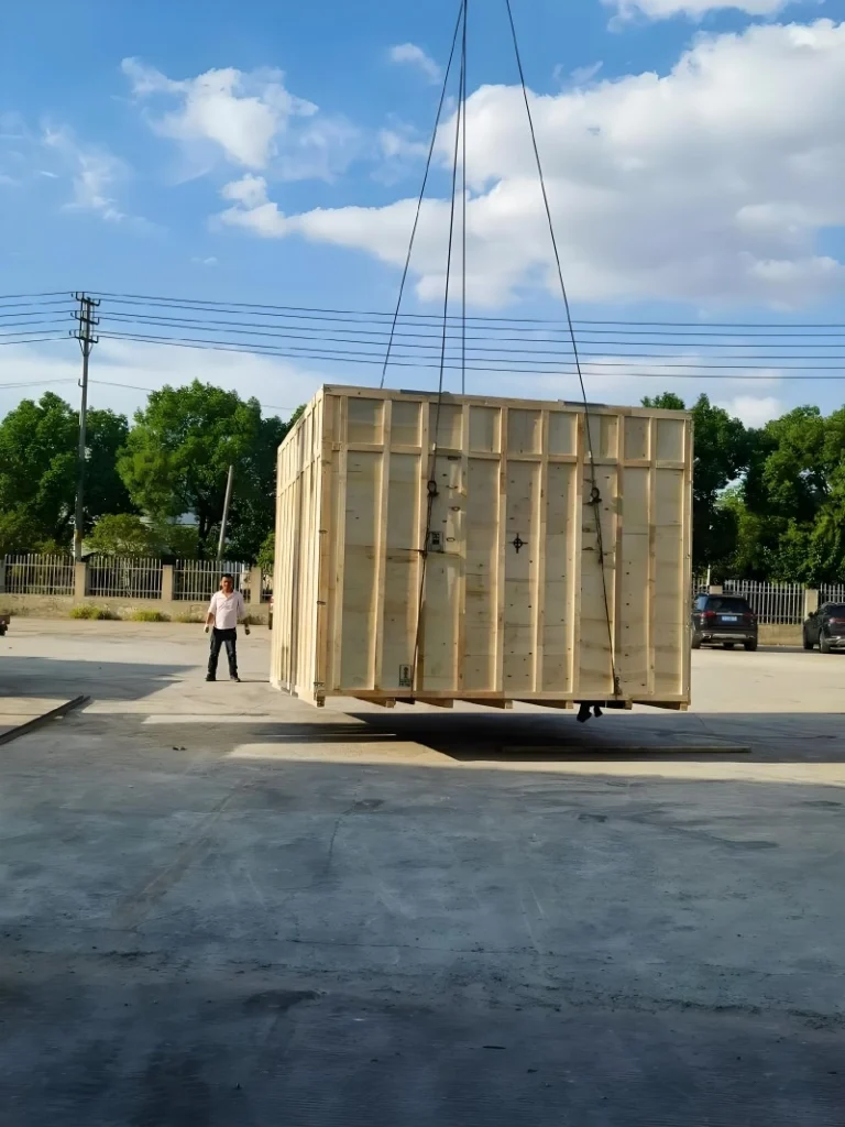 A large wooden export crate being lifted by a crane in an outdoor yard, with a worker guiding the load, demonstrating safe handling of heavy industrial equipment during road transport.